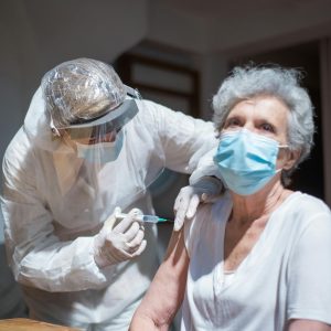 Elderly woman receiving a vaccine from a healthcare worker in protective gear indoors.
