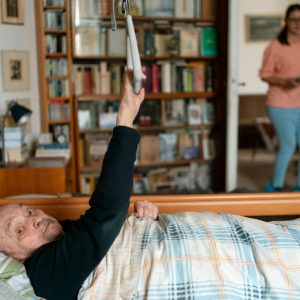 Senior man in bed with caregiver in Prague home, surrounded by books.
